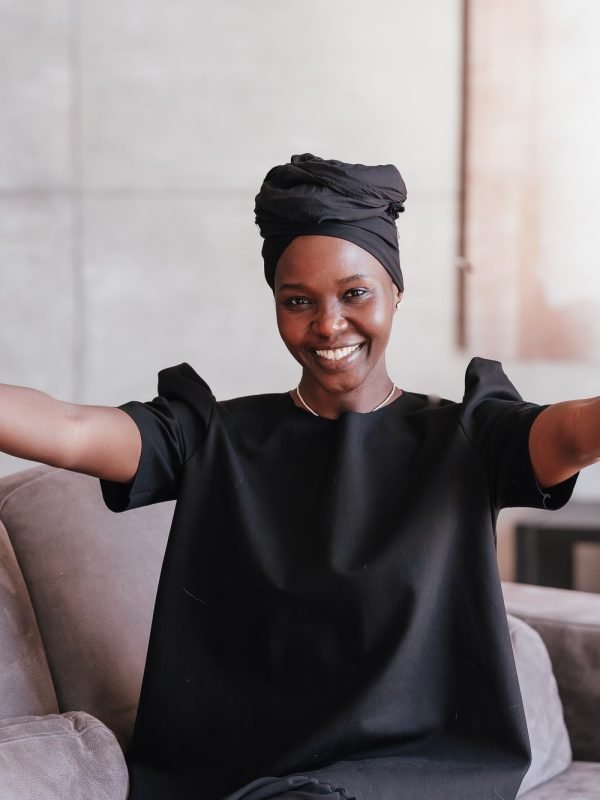 Cheerful African girl in black turban spreads hands looks at camera welcoming to hug sits on couch